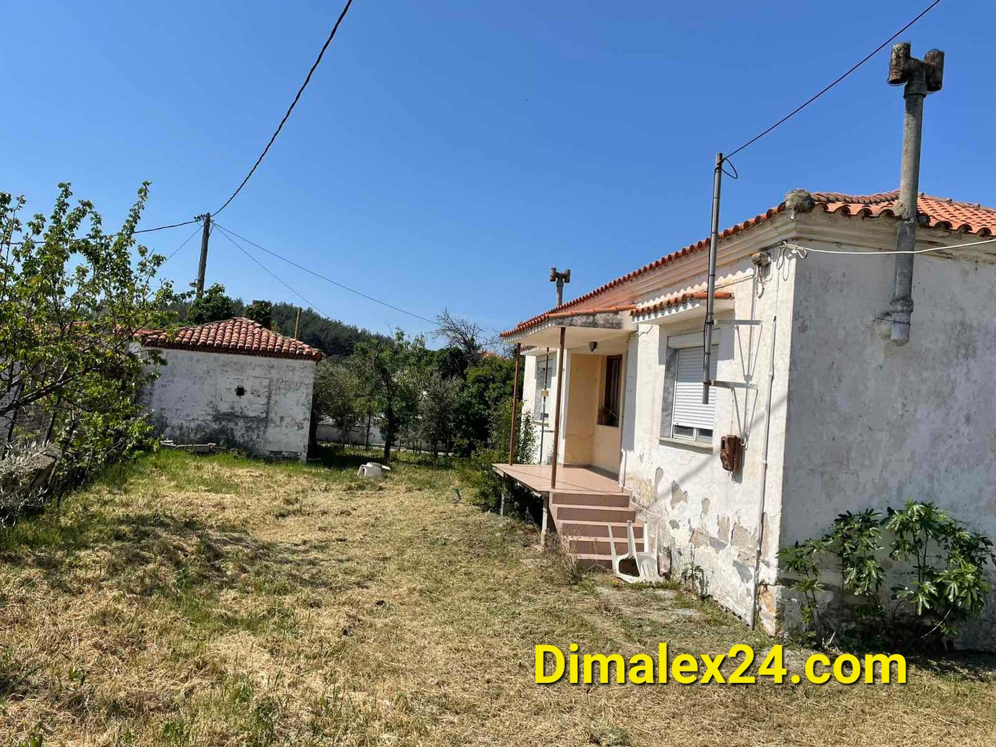 Exterior view of a house in Greece, featuring a garden and adjacent buildings, ideal for renovation projects.