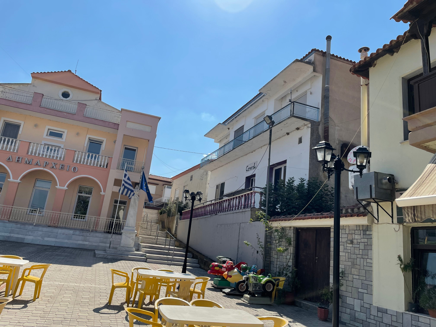 View of a sunny square in Ксилагани with colorful tables and buildings, showcasing local architecture.