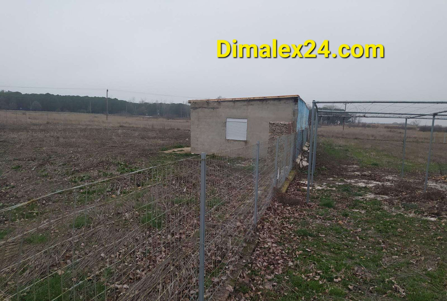 Small storage building surrounded by fencing on open land with sparse vegetation.