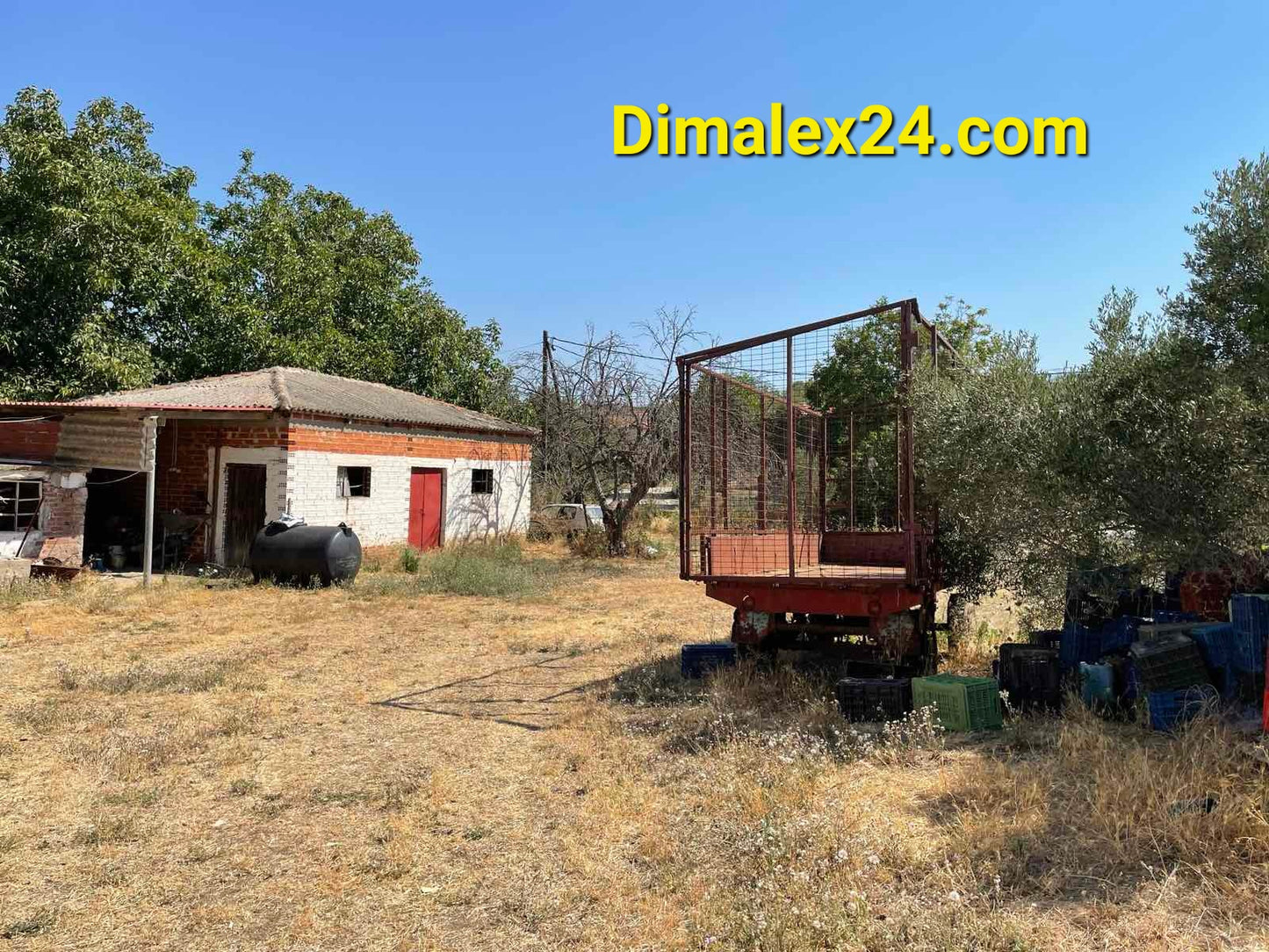 Old agricultural shed and equipment in a rural area with trees and dry grass.