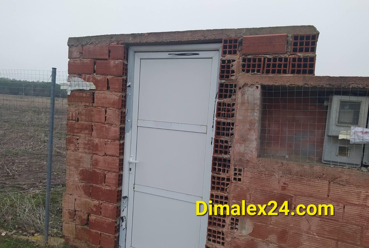 White door of a brick building in an open field, featuring a simple design and surrounding fencing.