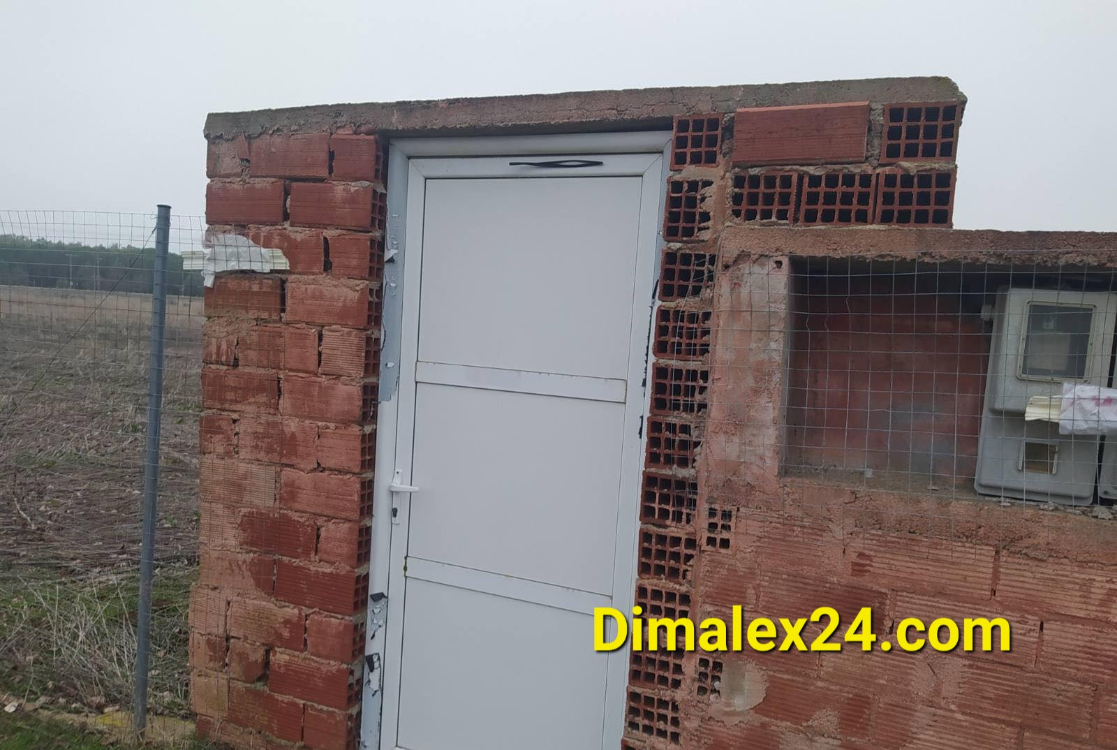 White door of a brick building in an open field, featuring a simple design and surrounding fencing.