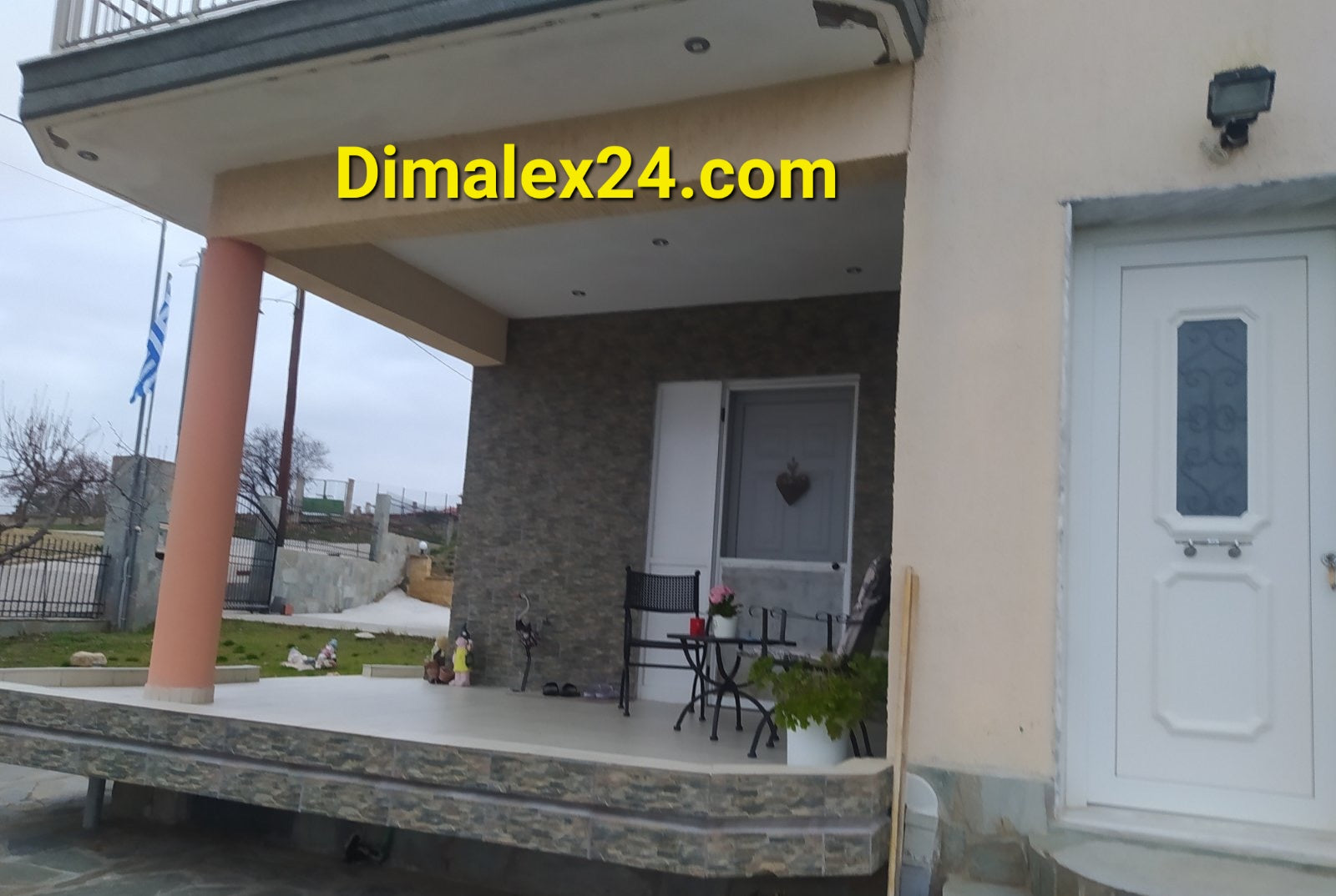 Entrance of a two-story house in Ksilo Gani, Northern Greece, featuring a patio and two chairs.