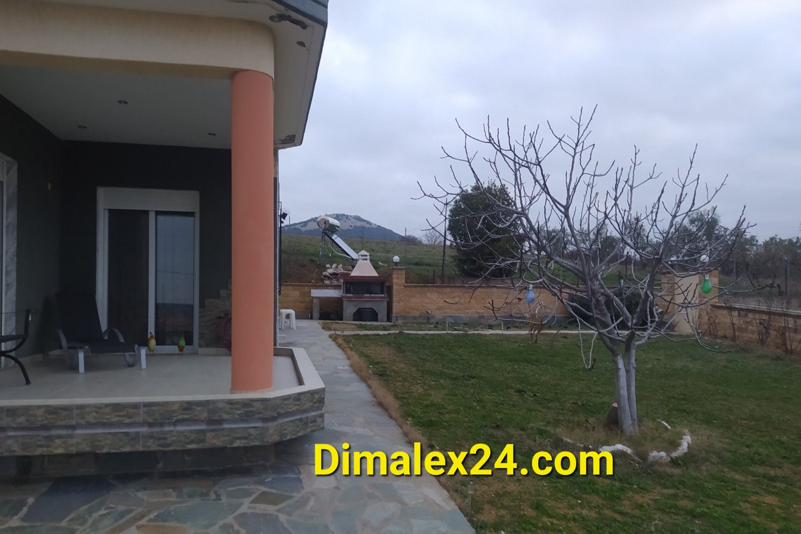 Exterior view of a two-story house in Northern Greece, featuring porch, garden, and barbecue area.