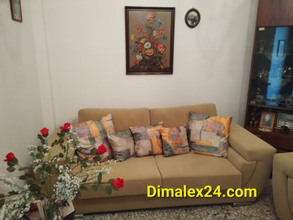 Cozy living room featuring a beige sofa with colorful cushions, floral wall art, and decorative plants.