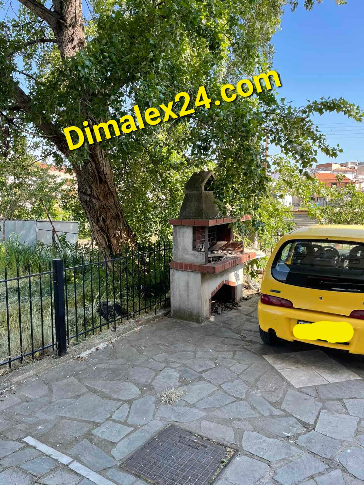 Yellow car parked near a barbecue area with a large tree and green surroundings in a residential area.